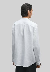 White linen long-sleeve shirt features a back yoke, side slits, and button cuffs, with a relaxed, slightly oversized fit.