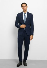 Marineblauer Anzug mit tailliertem Blazer und zulaufender Hose aus glattem Stoff. Kombiniert mit hellblauem Hemd und gemusterter Krawatte. Schwarze Schuhe.