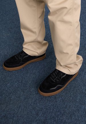 Fekete velúr sportcipő perforált részletekkel, fekete fűzőkkel és barna gumitalppal, bézs nadrággal párosítva egy kék szőnyegen.