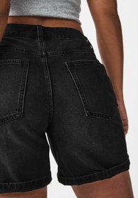 Pantalones cortos de denim negro con una cintura alta, dobladillos enrollados y dos bolsillos traseros con detalles de costura visibles. Textura suave y diseño ajustado.