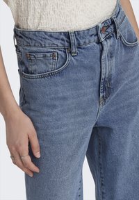 Höga jeans i blå denim med en texturerad yta. Funktioner inkluderar framfickor, dragkedja samt knoppar i kopparfärg.