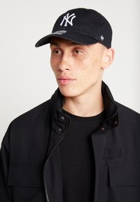 Black cap with white NY logo, cotton material, curved brim, and adjustable strap. Worn with a black zip-up jacket and t-shirt.