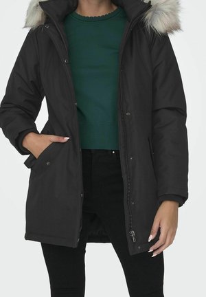 Femme portant un manteau d'hiver noir avec une capuche bordée de fourrure, un pull vert et un pantalon noir, main dans la poche du manteau, debout devant un fond uni.