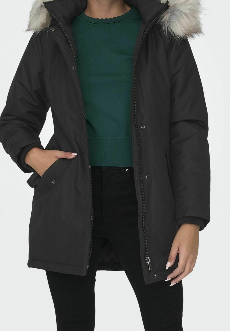 Femme portant un manteau d'hiver noir avec une capuche bordée de fourrure, un pull vert et un pantalon noir, main dans la poche du manteau, debout devant un fond uni.