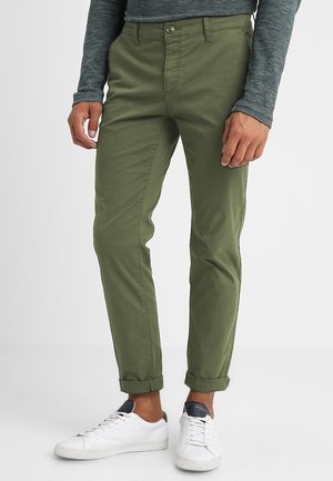 Chinos tapered de couleur vert khaki avec des ourlets roulés, en tissu de coton lisse. Portés avec des baskets blanches pour un look décontracté.