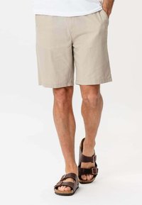 Mand iført beige knælange shorts og mørkebrune sandaler med dobbeltrem, der står med den ene hånd i lommen på hvid baggrund.