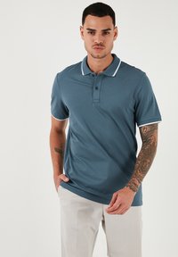 Teal polo majica iz gladkega materiala, s klasičnim ovratnikom z belim robom, dvema gumboma in kratkimi rokavi.