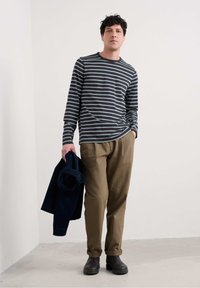 Viimeistelty tummanharmaa ja vaaleansininen raidallinen pitkähihaisen paidan yhdistelmä, joka on yhdistetty beigen värisiin kapeneviin housuihin. Pitelee tummansinistä takkia, jalassa ruskeat kengät.