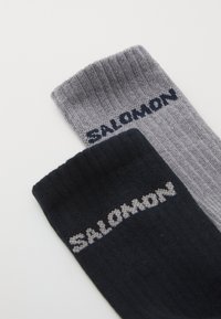 Δύο ζεύγη κάλτσες σε γκρι και σκούρο ναυτικό, με ραβδωτή υφή και λογότυπο "SALOMON" σε αντίθετα χρώματα.