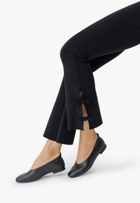 Jalad, millel on mustad püksid külgmiste lõhede ja väikeste paeltega, koos mustade nahast lamedate kingadega valgel taustal.