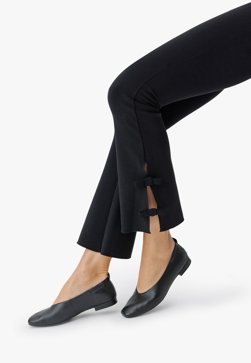 Jalad, millel on mustad püksid külgmiste lõhede ja väikeste paeltega, koos mustade nahast lamedate kingadega valgel taustal.