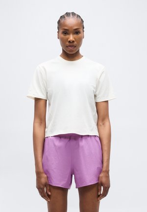 Vrouw met gevlochten haar, gekleed in een eenvoudig wit T-shirt met korte mouwen en lavendelkleurige sportshorts, staand tegen een effen lichte achtergrond.