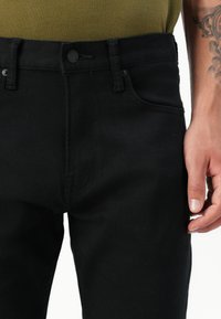 Juodos džinsinės kelnės su klasikinio kirpimo, turinčios mygtuką, šonines kišenes ir lygų paviršių. Diržo linija turi subtilų dizaino elementą.