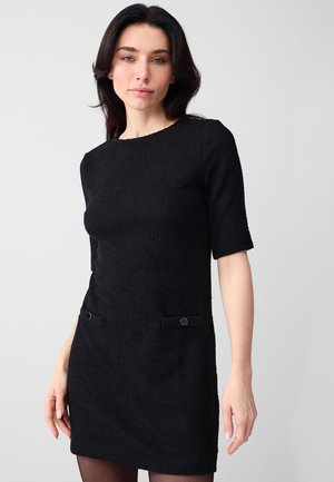 Kobieta o ciemnych włosach w teksturowanej czarnej sukience z krótkim rękawem i zapinanymi na guziki kieszeniami z przodu, stoi na tle jasnego, jednolitego tła.