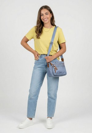 Nuori nainen hymyilee, pukeutuneena keltaiseen t-paitaan, vaalean sinisiin farkkuihin, valkoisiin lenkkareihin ja kantaa vaaleansinistä olkalaukkua, jossa on useita vetoketjullisia taskuja.