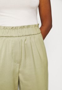 Pantalones verde claro con cintura fruncida, que cuentan con un solo bolsillo lateral y una textura lisa, combinados con una camiseta sin mangas blanca ajustada.