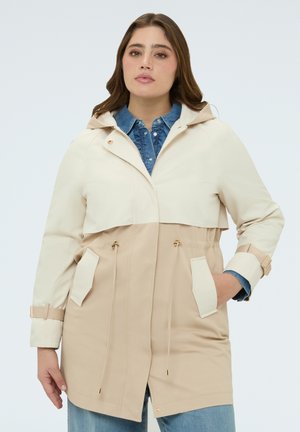 Kvinde iført beige og cremefarvet hættetrøje over blå denims skjorte med hænderne i jakkelommerne, stående foran en ensfarvet baggrund.