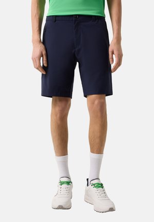 Mann trägt marineblaue Shorts, weiße Socken, weiße Turnschuhe mit grünen Schnürsenkeln und ein grünes Hemd, steht vor einem schlichten Hintergrund.