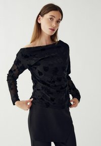 Hosszú hajú nő áttetsző, fekete virágmintás, hosszú ujjú blúzt visel lecsúszott vállú nyakkivágással, fekete szoknyával, fehér háttér előtt.