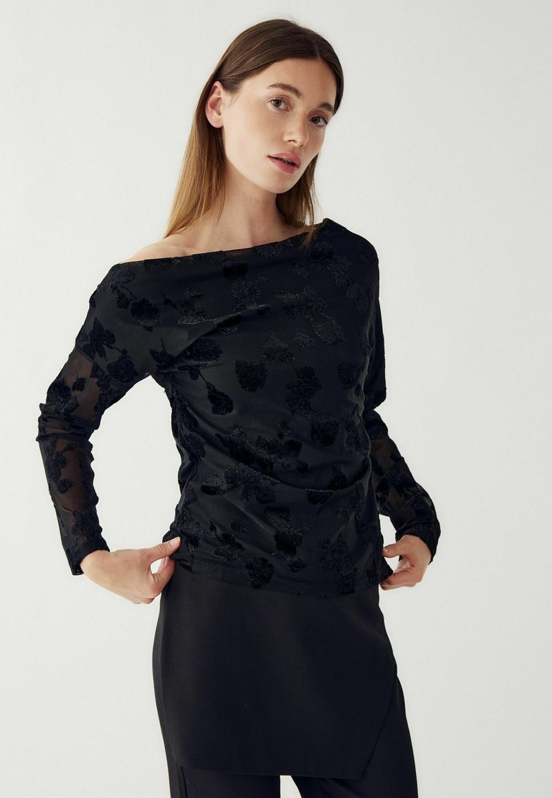 Hosszú hajú nő áttetsző, fekete virágmintás, hosszú ujjú blúzt visel lecsúszott vállú nyakkivágással, fekete szoknyával, fehér háttér előtt.