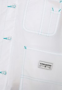Camicia bianca con bottoni, cuciture turchesi, una tasca sul petto e un'etichetta Converse in bianco e nero. Il tessuto ha un aspetto testurizzato.