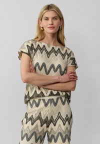 Beige top en broek set met een golvend zwart en beige patroon. Korte mouwen, losse pasvorm, textiel met structuur voor extra dimensie. Gouden creolen.
