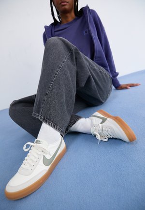 Bele tene Nike športne copate z sivimi detajli, z gumijastim podplatom, obuti z belimi nogavicami. Temno sivi, rahlo uničeni široki kavbojki. Modra ozadja.