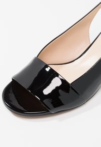 Chaussure plate à bout ouvert en cuir verni noir, avec une large bride à l'avant et une doublure intérieure beige sur fond blanc.