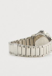 Silver metal link watch band with a clasp featuring a rectangular logo, clasp closed and curved around on a white background.