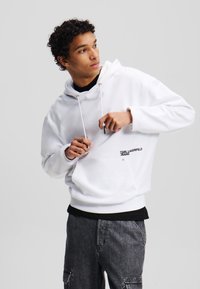 Weißer Hoodie mit einer vorderen Kängurutasche und schwarzem Logodetail. Verfügt über eine Kapuze mit Kordelzug, lange Ärmel und eine entspannte Passform. Kombiniert mit dunkelgrauen Hosen.