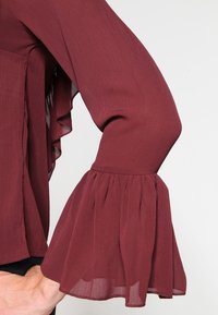 Bras droit plié, portant un haut à manches longues bordeaux avec des manches évasées à volants en tissu semi-transparent.