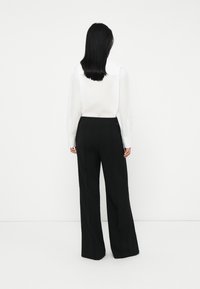 Person with long black hair wearing a white long-sleeve shirt and black wide-leg pants, standing facing a white wall.