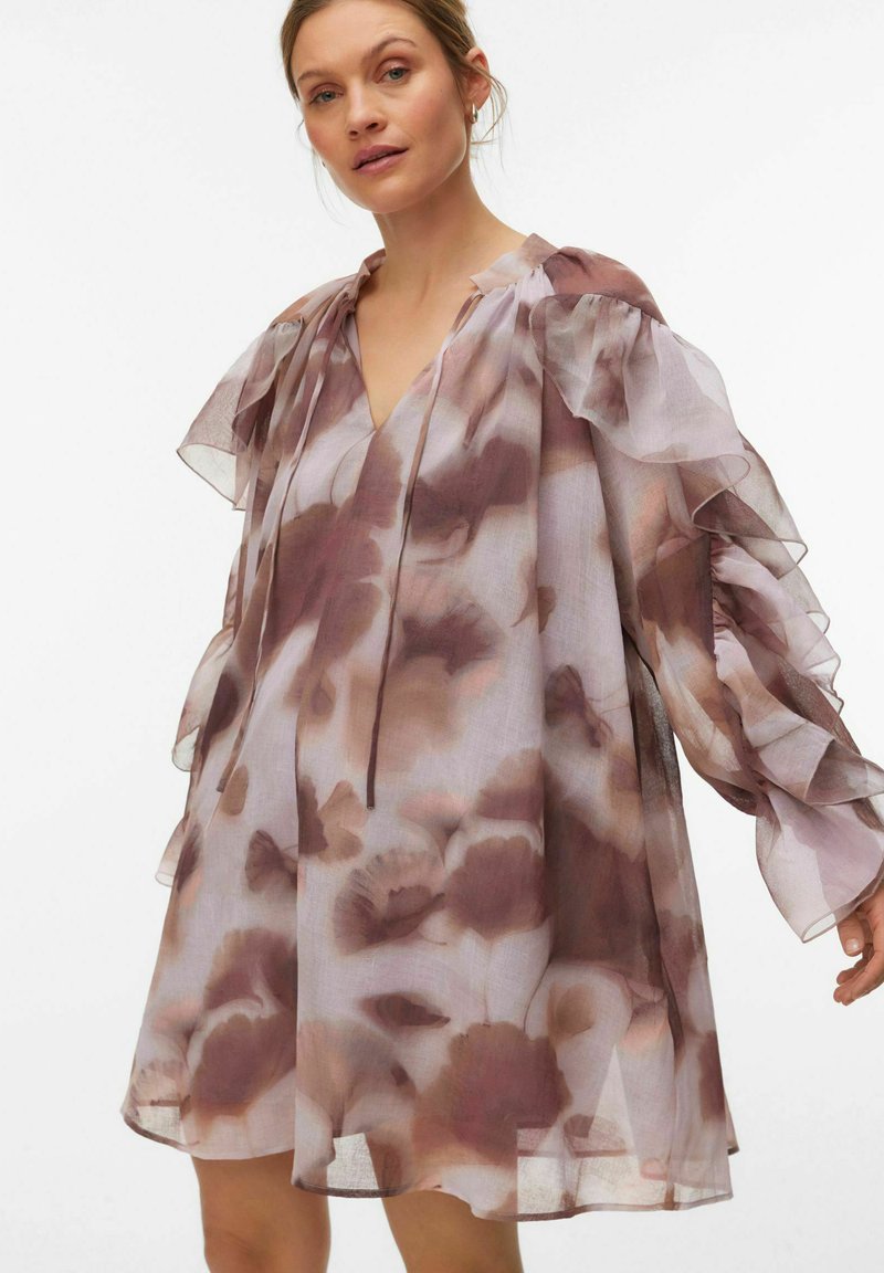 Vrouw draagt een losse, doorschijnende jurk met bruine en beige bloemenpatronen, voorzien van volumineuze gerimpelde mouwen en een V-hals met striksluiting.