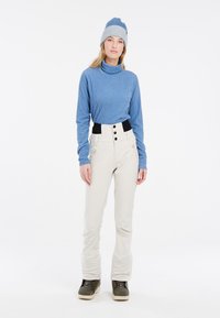 Blauer gerippter Rollkragenpullover, cremfarbene hochgeschneiderte Hose mit schwarzem Bund und Knöpfen, kombiniert mit einer grauen und blauen gestrickten Mütze und grünen Stiefeln.