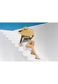 Cappello di paglia a larghe falde, top corto bianco, gonna bianca e sandali decorati. Il soggetto è seduto su gradini bianchi contro un cielo blu.