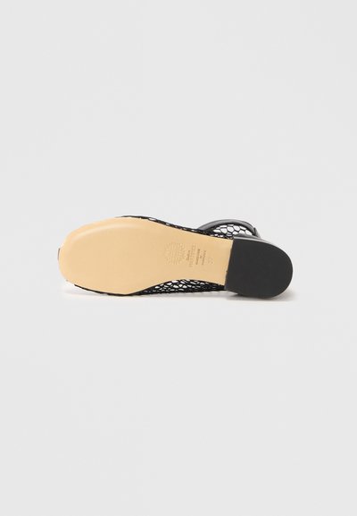 Sandal nera piatta con design a rete intrecciata e suola color beige chiaro, vista dalla parte inferiore su sfondo neutro.