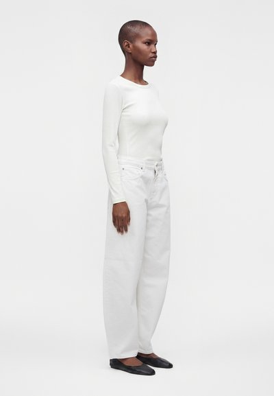 Personne de profil portant un haut blanc ajusté à manches longues, un jean blanc ample et des chaussures plates noires sur un fond blanc uni.