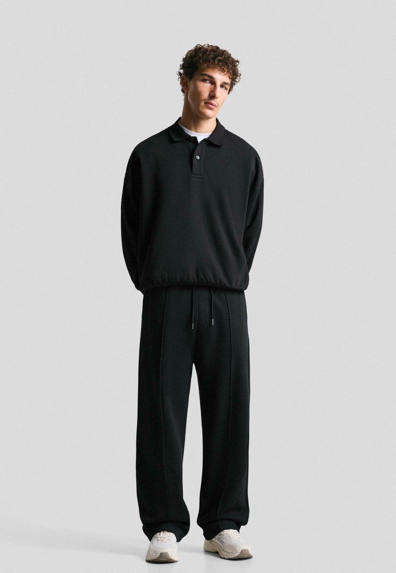 Black athletic outfit featuring a long-sleeve polo with a collar and button, loose-fit pants with drawstring, and light-colored shoes.