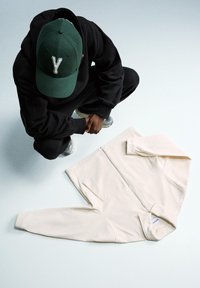 Cream-colored zip-up jacket with textured fabric, featuring a high collar and subtle stitching details. Black hoodie and green cap visible.