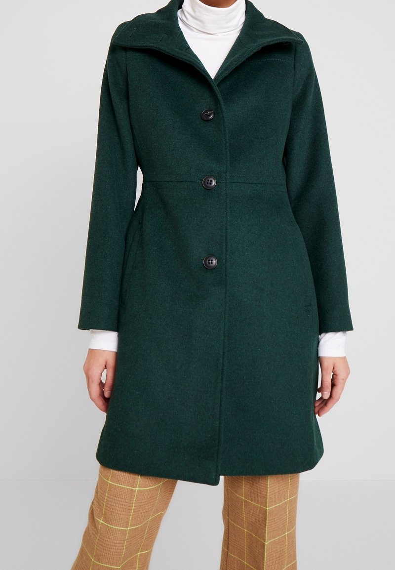 Femme portant un manteau vert foncé boutonné sur un col roulé blanc et un pantalon marron à carreaux, debout devant un fond uni.