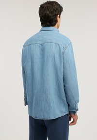 Hombre de espaldas vistiendo una camisa de mezclilla azul claro con puños abotonados y pantalones azul oscuro sobre un fondo liso.