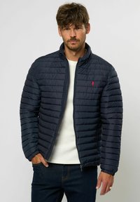 Marineblå quiltet jakke med stående krage, som har et diagonalt stikkmønster og en liten rød logo på brystet. Glatt, lett materiale.