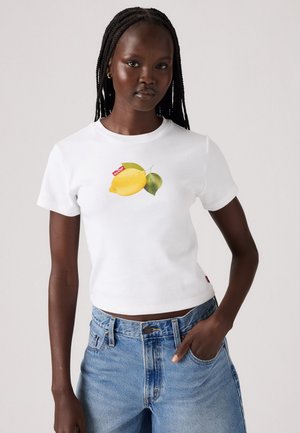 Biely bavlnený tričko s veľkým žltým grafickým motívom citróna so zelenými listami, krátkymi rukávmi a priliehavým strihom. Nosené s modrými džínsami.