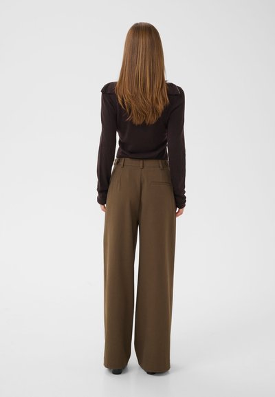 Femme aux longs cheveux bruns et lisses porte un haut noir à manches longues et un pantalon marron taille haute à jambes larges, debout de dos sur un fond blanc.