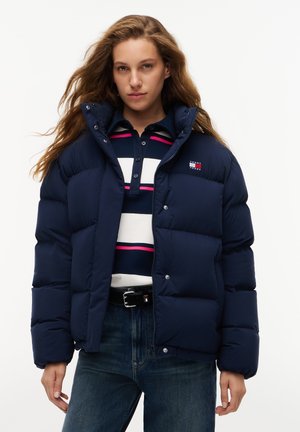 Frau trägt eine marineblaue Steppjacke über einem gestreiften Poloshirt, blaue Jeans und einen schwarzen Gürtel, steht vor einem weißen Hintergrund.