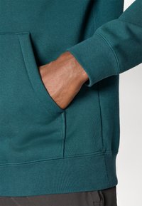 Mano di colore scuro parzialmente all'interno della tasca anteriore di una felpa color teal con polsini a coste e dettagli delle cuciture visibili.