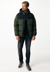 Grøn og sort pufferjakke med hætte, der har et quiltet design, elastiske manchetter og lynlås foran. Kombinationen er med mørke jeans og sneakers.