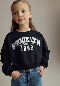 Sweatshirt noir avec "BROOKLYN UNIVERSITY LEAGUE 1982" imprimé en blanc, associé à un short en denim bleu clair avec des poches visibles.