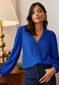 Modra bluzica z vzorcem, z V-izrezom, nabranimi rokavi in čipkastimi poudarki. Združena s temnim jeansa, z gumbom in žepnimi detajli.