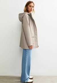 Veste imperméable beige avec capuche, fermeture à boutons sur le devant et poches plaquées, assortie à un jean ample bleu clair et des chaussures blanches.